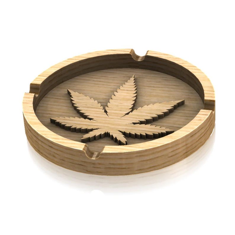 May include: A wooden ashtray featuring a carved marijuana leaf design. The ashtray is round and has a brown finish.