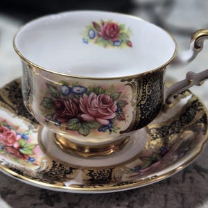 May include: A white teacup and saucer with a black and gold floral pattern. The cup has a gold rim and a delicate handle. The saucer has a matching pattern and a gold rim.