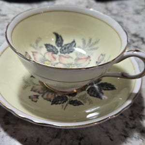 May include: A vintage teacup and saucer set. The cup and saucer are pale yellow with a floral design in pink, gray, and black. The rims are trimmed with gold. The cup has a curved handle with gold accents.