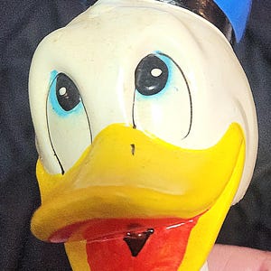 May include: A vintage ceramic figurine of Donald Duck wearing a blue sailor hat with a black band. The figurine has a yellow beak, red mouth, and blue eyes.