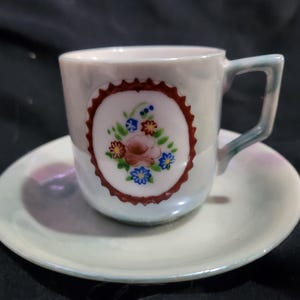 Puede incluir: Un juego de taza y platillo de té de porcelana blanca con un diseño floral. La taza tiene un borde rojo y un motivo floral en el centro. El platillo es blanco con un glaseado iridiscente rosa claro y azul.