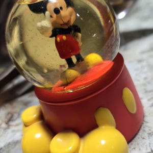 May include: A clear glass snow globe with a Mickey Mouse figurine inside. The base of the snow globe is red with yellow Mickey Mouse ears.