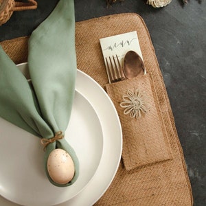 Può includere: Un tovagliolo verde piegato a forma di orecchie di coniglio con un uovo macchiato legato al centro, posto su un piatto bianco. Il piatto è su un sottopiatto di juta marrone con una tasca che contiene un menu, posate e un fiore bianco.