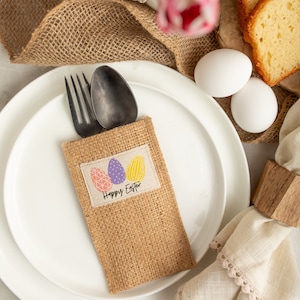 Könnte beinhalten: Ein Serviettenhalter aus Jute mit einem "Frohe Ostern"-Design mit drei bunten Ostereiern. Der Halter befindet sich auf einem weißen Teller mit einer Gabel und einem Löffel darin.