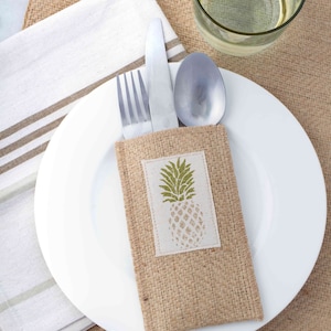 May include: A burlap cutlery holder with a pineapple design. The holder is brown and has a white fabric patch with a green pineapple on it. The holder is holding a fork, knife, and spoon.