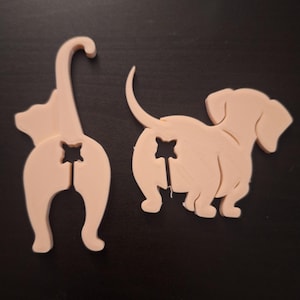 May include: Two light beige animal-shaped cutouts. One is a cat, the other a dachshund dog. Both have a star-shaped cutout in the rear. The cat is standing, and the dog is sitting. The objects are on a dark surface.
