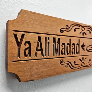 May include: A wooden sign with the text "Ya Ali Madad" carved into it. The sign has a decorative border with swirls and a star.