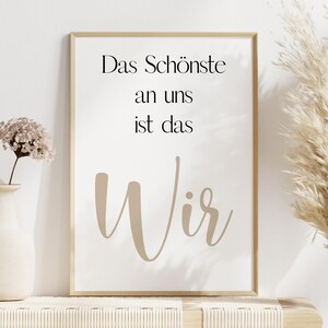 Puede incluir: Una impresión enmarcada con el texto alemán "Das Schönste an uns ist das Wir" en negro y beige. La impresión se muestra en una pared blanca, con jarrones decorativos y hierba de la pampa en el fondo.