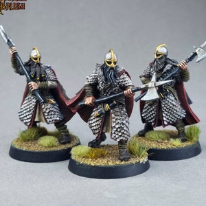 May include: Three painted miniature figures of warriors wearing chainmail armor, horned helmets, and red cloaks. Each figure holds a weapon, including an axe and a spear. The figures are standing on circular bases with a grassy texture.