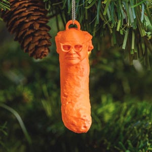 May include: Orange novelty Christmas ornament shaped like a pickle with a face and glasses. The ornament is hanging from a silver chain against a backdrop of green Christmas tree branches and a pine cone.