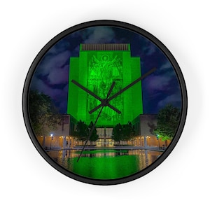 Könnte beinhalten: Eine runde Wanduhr mit schwarzem Rahmen. Das Zifferblatt zeigt ein grün beleuchtetes Gebäude mit einem Wandbild vor einem Nachthimmel. Schwarze Zeiger zeigen die Uhrzeit an.