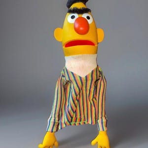 May include: A yellow puppet with a red nose, black hair, and a striped shirt. The puppet has yellow hands and a white turtleneck. The shirt has vertical stripes in various colors.