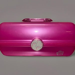 May include: A bright pink, rectangular makeup case with a clear handle and a circular emblem that reads "STYLE". The case has a glossy finish and a small logo on the bottom right corner.