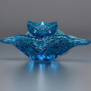 Puede incluir: Un juego de dos cuencos decorativos de cristal azul. El cuenco más grande tiene un borde festoneado y un diseño de estrella. El cuenco más pequeño se encuentra encima del más grande. El cristal tiene un diseño de cristal tallado.