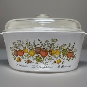May include: White casserole dish with a clear glass lid. The dish is decorated with a colourful vegetable and herb design, including tomatoes, lemons, and herbs. The text "L'échalote, La Marjolaine, Le Romarin" is printed on the front.