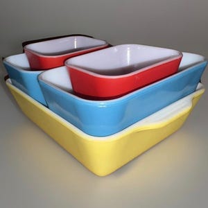 Op de afbeelding: Een stapel kleurrijke rechthoekige bakvormen. De set bevat een gele basis, een blauwe schaal, een rode schaal en een zwarte schaal. Elke schaal heeft een wit interieur. De schalen zijn in elkaar genest.