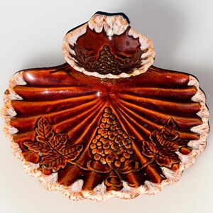 Puede incluir: Un plato decorativo de cerámica marrón con borde festoneado y diseño de hojas. El plato presenta un racimo de uvas central y detalles de hojas, con un plato más pequeño y elevado en la parte superior. El borde es blanco y marrón.