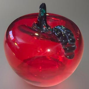 May include: A vibrant red glass apple sculpture with a dark blue stem and a touch of green. The glossy surface reflects light, creating a striking decorative piece. This handcrafted art object is a unique home decor item.