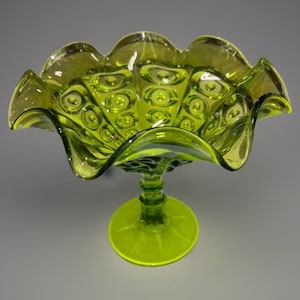 Könnte beinhalten: Eine grüne Glas-Schale mit Fuß und gewelltem Rand. Die Schale hat ein strukturiertes Muster und steht auf einem runden Sockel. Das Glas hat eine lebendige, durchscheinende Qualität.