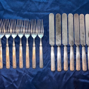 Op de afbeelding: Een set antiek zilverwerk, inclusief vorken en messen, gerangschikt op een blauw oppervlak. De vorken hebben vier tanden en sierlijke handvatten. De messen hebben afgeronde bladen en bijpassende handvatten, wat wijst op een complete eetservies.