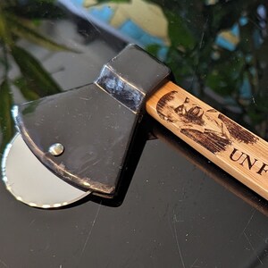 May include: A black pizza cutter shaped like an axe with a wooden handle. The handle is engraved with the text "UN FORASTERO!!"