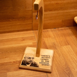 Puede incluir: Un soporte de plátanos de madera con una parte superior curvada y una base rectangular. La base presenta un retrato de un hombre y el texto "THERE'S ALWAYS MONEY IN THE BANANA STAND".