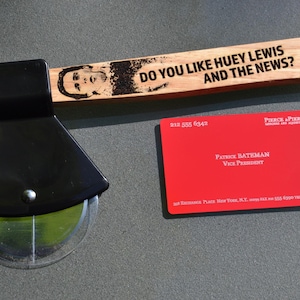 Puede incluir: Un cortador de pizza de madera con un mango negro y una rueda de plástico transparente. El mango tiene el texto "Do you like Huey Lewis and the News?" grabado. También se ve una tarjeta de visita roja con el texto "Patrice Bateman Vice President" en la imagen.