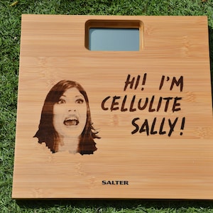 May include: A wooden bathroom scale with a digital display and a humorous message that reads "Hi! I'm Cellulite Sally!" The scale is made by Salter.