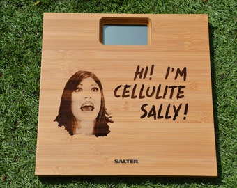 May include: A wooden bathroom scale with a digital display and a humorous message that reads "Hi! I'm Cellulite Sally!" The scale is made by Salter.