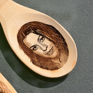 Puede incluir: Una cuchara de madera con una imagen grabada con l&aacute;ser de una cara. La imagen est&aacute; en tonos marrones y negros.