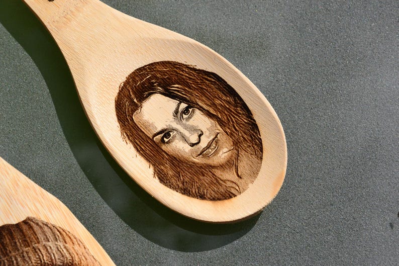 Puede incluir: Una cuchara de madera con una imagen grabada con l&aacute;ser de una cara. La imagen est&aacute; en tonos marrones y negros.