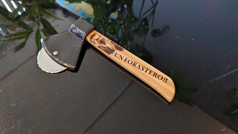 May include: A black pizza cutter shaped like an axe with a wooden handle. The handle is engraved with the text "UN FORASTERO!!"