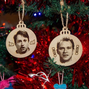 Peut inclure: Deux décorations de Noël en bois suspendues à un sapin décoré. Chaque ornement rond présente un portrait d'une personne. L'un a une expression surprise et des images de dinde, l'autre a une expression neutre et de petites boîtes.