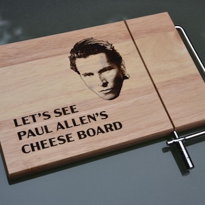 Puede incluir: Tabla de queso de madera con retrato y texto. La tabla marrón claro presenta un retrato y las palabras "LET'S SEE PAUL ALLEN'S CHEESE BOARD" en negro. Un cortador de queso de metal está adjunto.