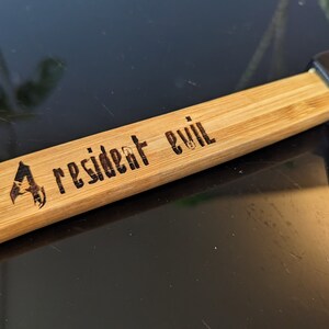 May include: A wooden axe handle with a black axe head. The handle is engraved with the words "Resident Evil" and a logo.