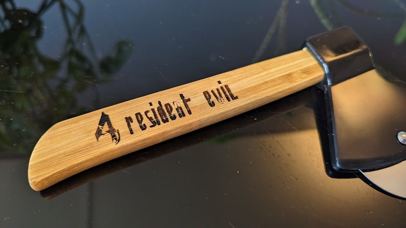 May include: A wooden axe handle with a black axe head. The handle is engraved with the words "Resident Evil" and a logo.