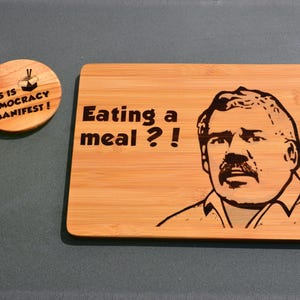 Puede incluir: Una tabla de cortar de madera con una imagen grabada con láser de un hombre con bigote y el texto "¿Comiendo una comida?!". La tabla es de color marrón claro y tiene una veta de madera natural. Hay un pequeño posavasos redondo de madera con el texto "¡Este es el Manifiesto de la Democracia!" y una pequeña imagen de un contenedor para llevar con palillos.