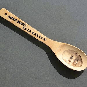 May include: A wooden spoon with the text "ARNE SLOT! LA LA LA LA LA!" etched in black. The spoon's bowl features a detailed engraved portrait of a smiling man. The spoon is made of light-coloured wood.
