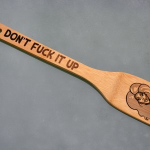 Könnte beinhalten: Bambus-Pfannenwender mit dem eingebrannten Schriftzug "DON'T FUCK IT UP" auf dem Griff. Der Pfannenwender zeigt eine Cartoon-Illustration eines Frauengesichts mit langen Haaren. Der Pfannenwender hat oben ein Loch.