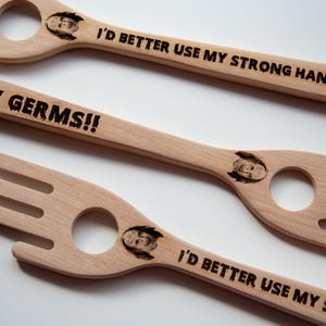 Puede incluir: Tres utensilios de ensalada de madera con un extremo en forma de tenedor y un agujero redondo. El texto "MY GERMS!!" está grabado en un utensilio, mientras que los otros dicen "I'D BETTER USE MY STRONG HAND". Cada utensilio tiene una pequeña cara grabada.