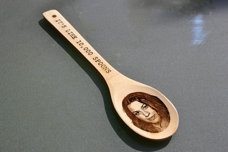 Puede incluir: Una cuchara de madera con el texto "It's like 10,000 spoons" grabado en el mango. Un rostro de mujer est&aacute; grabado en el cuenco de la cuchara.
