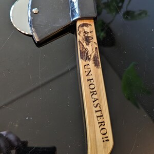 May include: A black and silver pizza cutter shaped like an axe with a wooden handle. The handle has a burned-in image of a man's face and the text "UN FORASTER0!!"