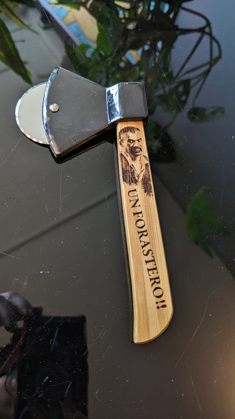 May include: A black and silver pizza cutter shaped like an axe with a wooden handle. The handle has a burned-in image of a man's face and the text "UN FORASTER0!!"
