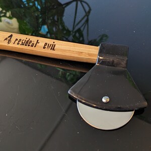 May include: A black pizza cutter shaped like an axe with a wooden handle. The handle is engraved with the words "Resident Evil".