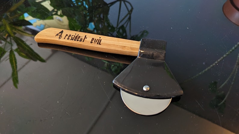 May include: A black pizza cutter shaped like an axe with a wooden handle. The handle is engraved with the words "Resident Evil".