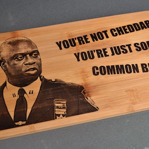 Könnte beinhalten: Rechteckiges Schneidebrett aus hellem Holz mit einem lasergravierten Bild eines Mannes in Polizeiuniform. Der Text "YOU'RE NOT CHEDDAR YOU'RE JUST SOME COMMON BITCH" befindet sich über dem Bild.