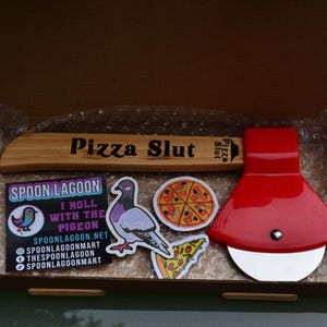 May include: A pizza-themed gift set in a cardboard box. Includes a wooden pizza cutter with "Pizza Slut" engraved, a red pizza wheel, and several stickers featuring a pigeon and pizza slices. The stickers also include the text "Spoon Lagoon".