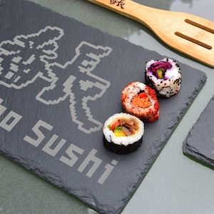 May include: Two black slate serving trays with the text "Bancho Sushi" and a pixelated image of a Japanese character. The trays are on a table with a wooden spatula and three pieces of sushi.