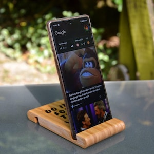 May include: A wooden phone stand with the text "DO IT FOR THE" engraved on it. A smartphone is propped up in the stand, displaying a news article about Donkey Kong.