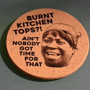 May include: A circular cork coaster featuring the text "BURNT KITCHEN TOPS?! AIN'T NOBODY GOT TIME FOR THAT" in black. A black and white image of a smiling woman is on the right. The coaster is brown.